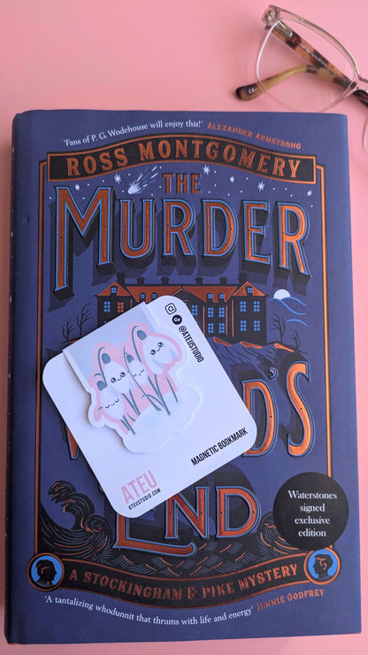 A Kawaii Sleepy Snowdrop 'Icy Venusian' pink magnetic bookmark on a branded card, angled to the left over a Waterstone's exclusive edition book. This aerial shot highlights the color contrast between the soft pink bookmark and the deep bronze lettering of the mystery novel, styled with elegant porcelain and glasses.