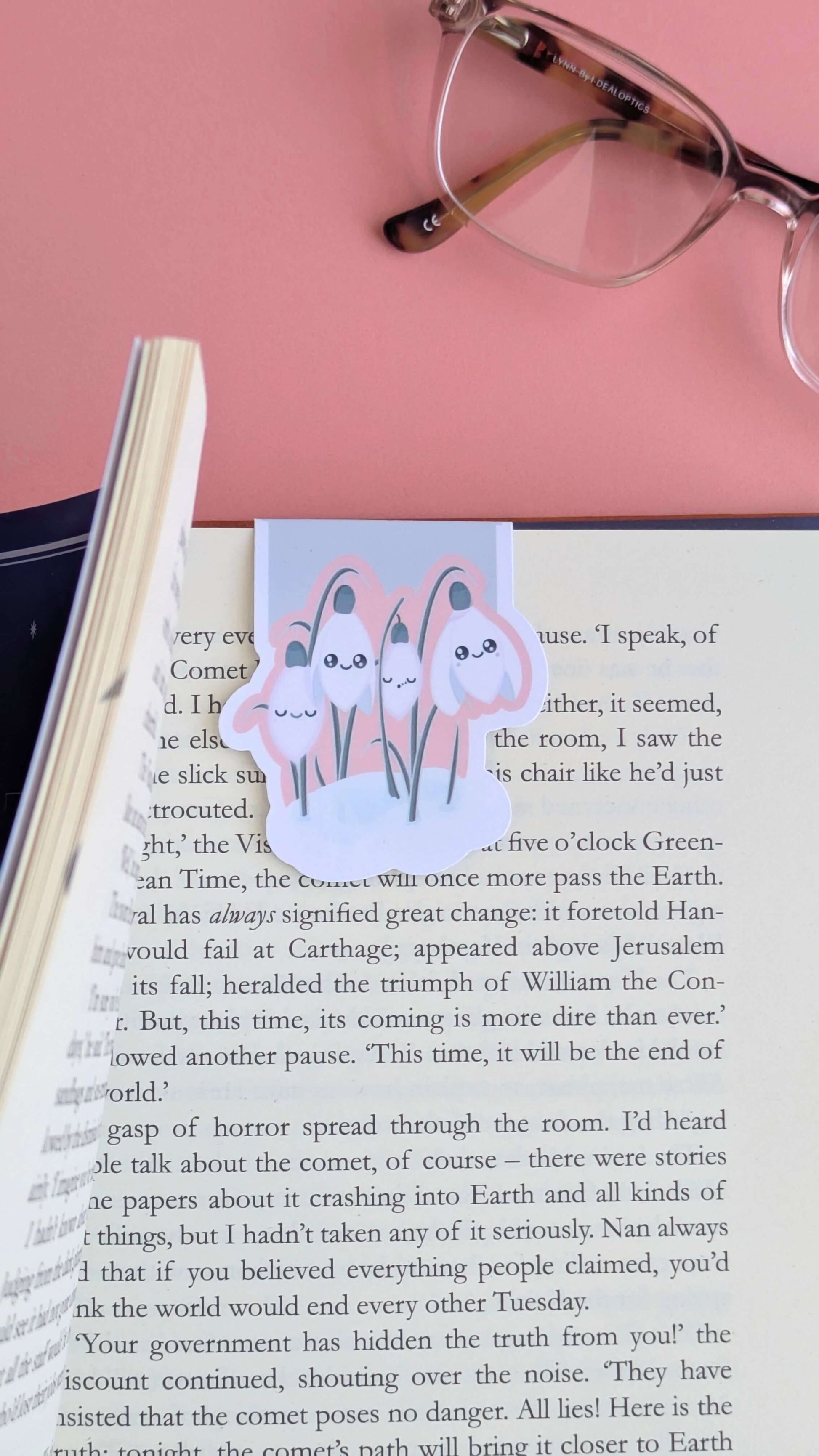 A close-up, intimate aerial view of an Ateu Studio Kawaii Sleepy Snowdrop magnetic bookmark being revealed for the first time as a book is opened. The left-hand pages are batched together, creating a sense of depth and movement, while the bookmark sits centrally on the page. Part of a pair of reading glasses and a soft pink background frame the top third of the image, capturing a quiet, aesthetic moment of a daily reading habit in natural light.