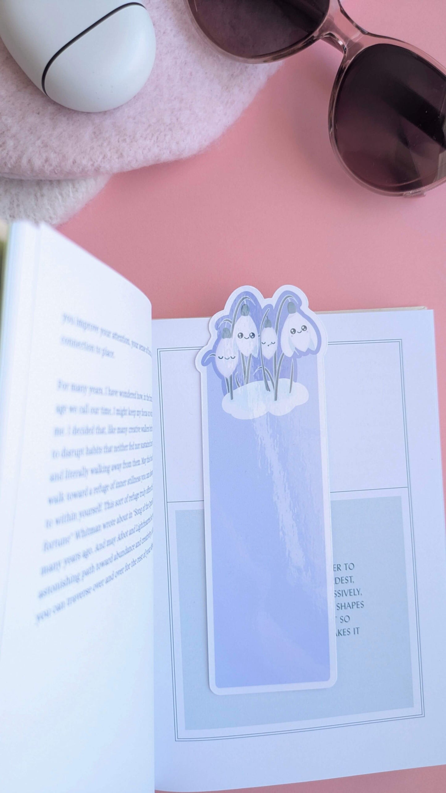 A Kawaii Sleepy Snowdrop 'Midnight Mist' bookmark centered on the title page of a book, where the silver-sage text echos the bookmark's foliage. The scene is styled on a soft pink winter hat with translucent taupe Fossil-style sunglasses on a rose-pink background. A long gloss sheen highlight reflects the light across the die-cut silhouette, showcasing a moody, romanticized reading ritual.