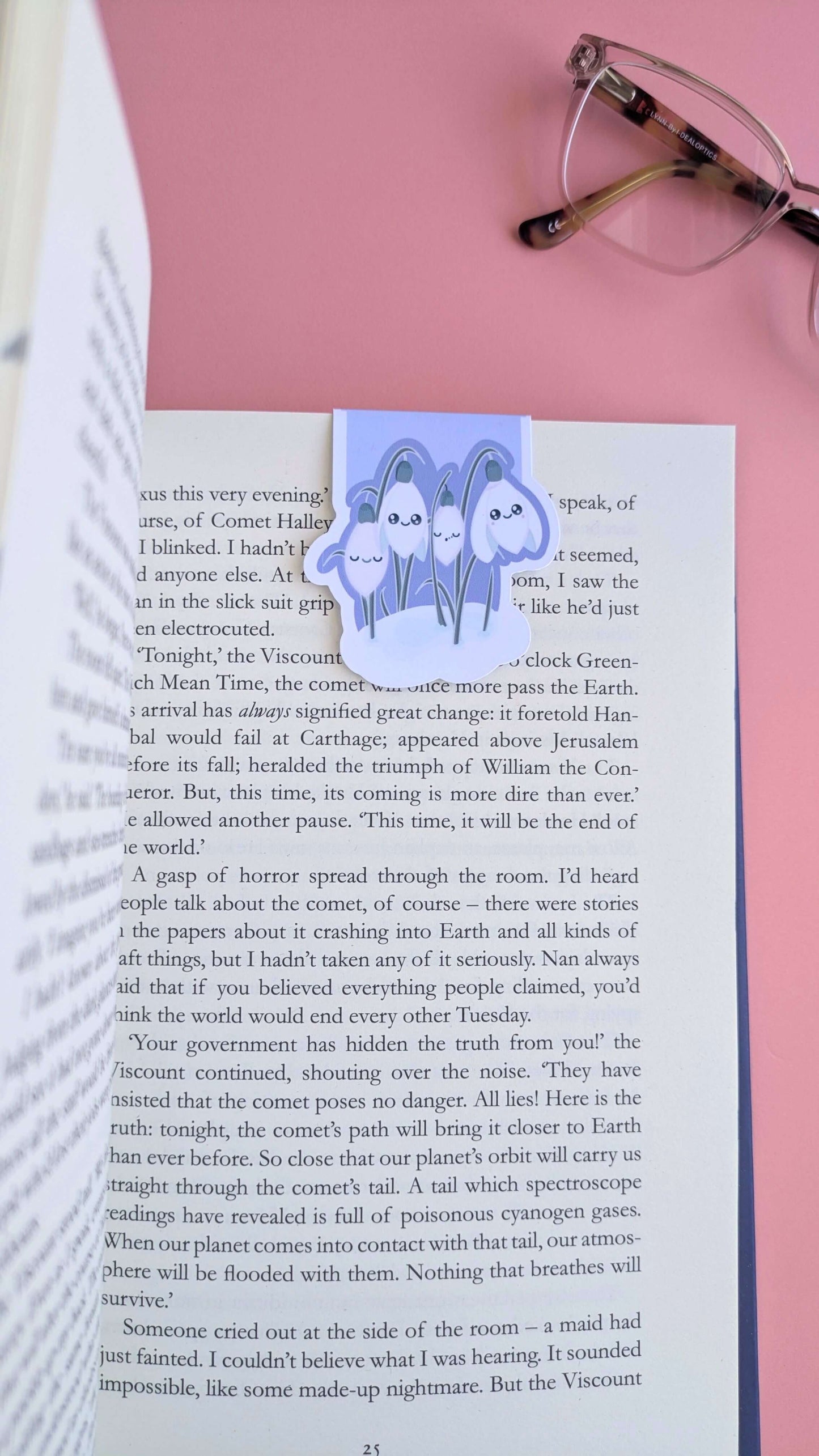 An aerial perspective looking down the right-hand page of an open book, featuring the Kawaii Sleepy Snowdrop 'Midnight Mist' lavender blue magnetic bookmark. The bookmark is positioned toward the right edge, styled with a single lens of a pair of reading glasses on a rose-pink background. The composition highlights the deep lavender tones against the printed page, capturing a natural-light snapshot of a focused, aesthetic reading session.