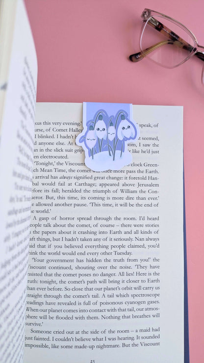 An aerial perspective looking down the right-hand page of an open book, featuring the Kawaii Sleepy Snowdrop 'Midnight Mist' lavender blue magnetic bookmark. The bookmark is positioned toward the right edge, styled with a single lens of a pair of reading glasses on a rose-pink background. The composition highlights the deep lavender tones against the printed page, capturing a natural-light snapshot of a focused, aesthetic reading session.
