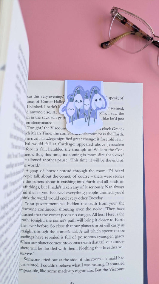 An aerial perspective looking down the right-hand page of an open book, featuring the Kawaii Sleepy Snowdrop 'Midnight Mist' lavender blue magnetic bookmark. The bookmark is positioned toward the right edge, styled with a single lens of a pair of reading glasses on a rose-pink background. The composition highlights the deep lavender tones against the printed page, capturing a natural-light snapshot of a focused, aesthetic reading session.