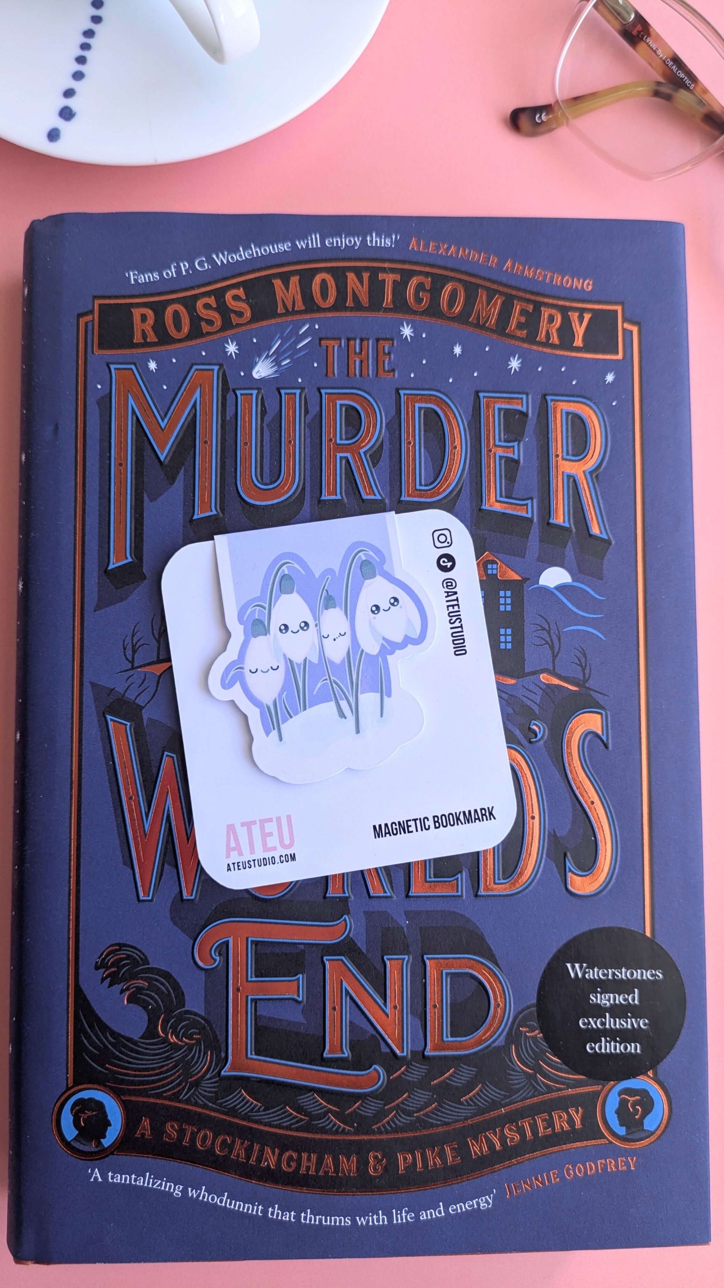 The Kawaii Sleepy Snowdrop 'Midnight Mist' lavender blue magnetic bookmark on its card, angled to the left. The deep blue tones of the bookmark harmonize with the starry night details of 'The Murder at World’s End' cover. Styled with a hand-painted porcelain cup and reading glasses on a pink background.