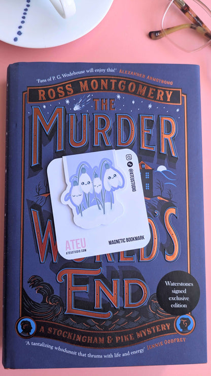 The Kawaii Sleepy Snowdrop 'Midnight Mist' lavender blue magnetic bookmark on its card, angled to the left. The deep blue tones of the bookmark harmonize with the starry night details of 'The Murder at World’s End' cover. Styled with a hand-painted porcelain cup and reading glasses on a pink background.