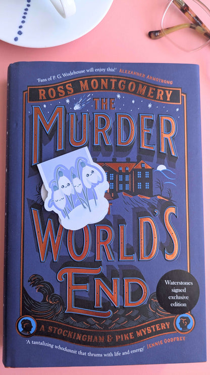 A Kawaii Sleepy Snowdrop 'Midnight Mist' magnetic bookmark angled to the left, adding to the atmospheric mood of the starry book cover. The composition features a porcelain saucer and reading glasses, emphasizing the bookmark as a high-quality accessory for mystery novel enthusiasts.