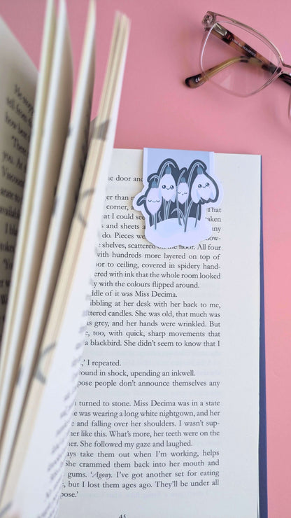 An aerial, close-up view of a book being opened to reveal a Kawaii Sleepy Snowdrop 'Midnight Mist' lavender blue magnetic bookmark holding a page. The left-hand pages are grouped and angled toward the lens, creating a sense of depth, while the right-hand page lies flat in natural daylight. Styled with the lens and bridge of reading glasses on a rose-pink background, this shot captures a mindful, aesthetic book annotating moment.