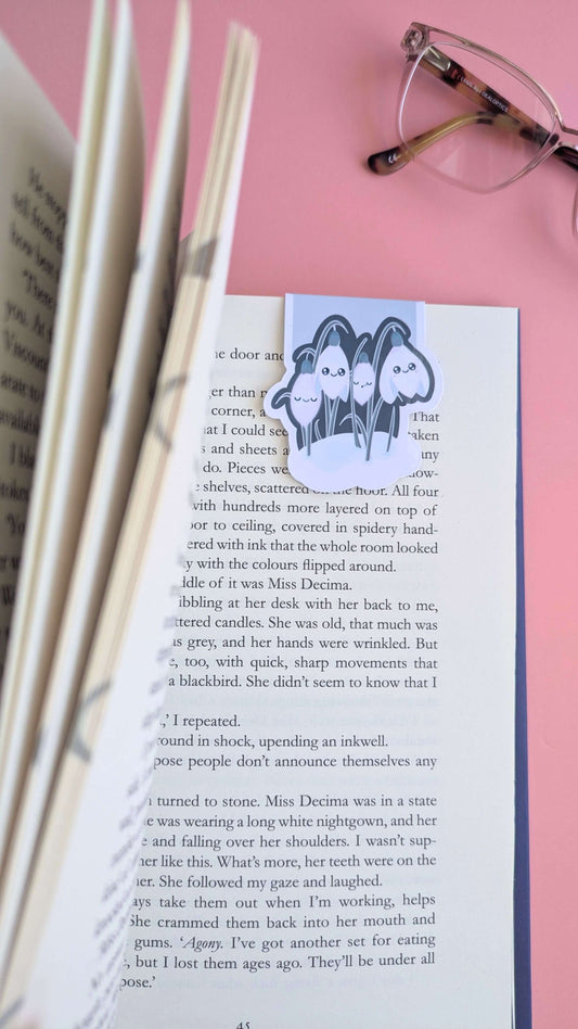 An aerial, close-up view of a book being opened to reveal a Kawaii Sleepy Snowdrop 'Midnight Mist' lavender blue magnetic bookmark holding a page. The left-hand pages are grouped and angled toward the lens, creating a sense of depth, while the right-hand page lies flat in natural daylight. Styled with the lens and bridge of reading glasses on a rose-pink background, this shot captures a mindful, aesthetic book annotating moment.