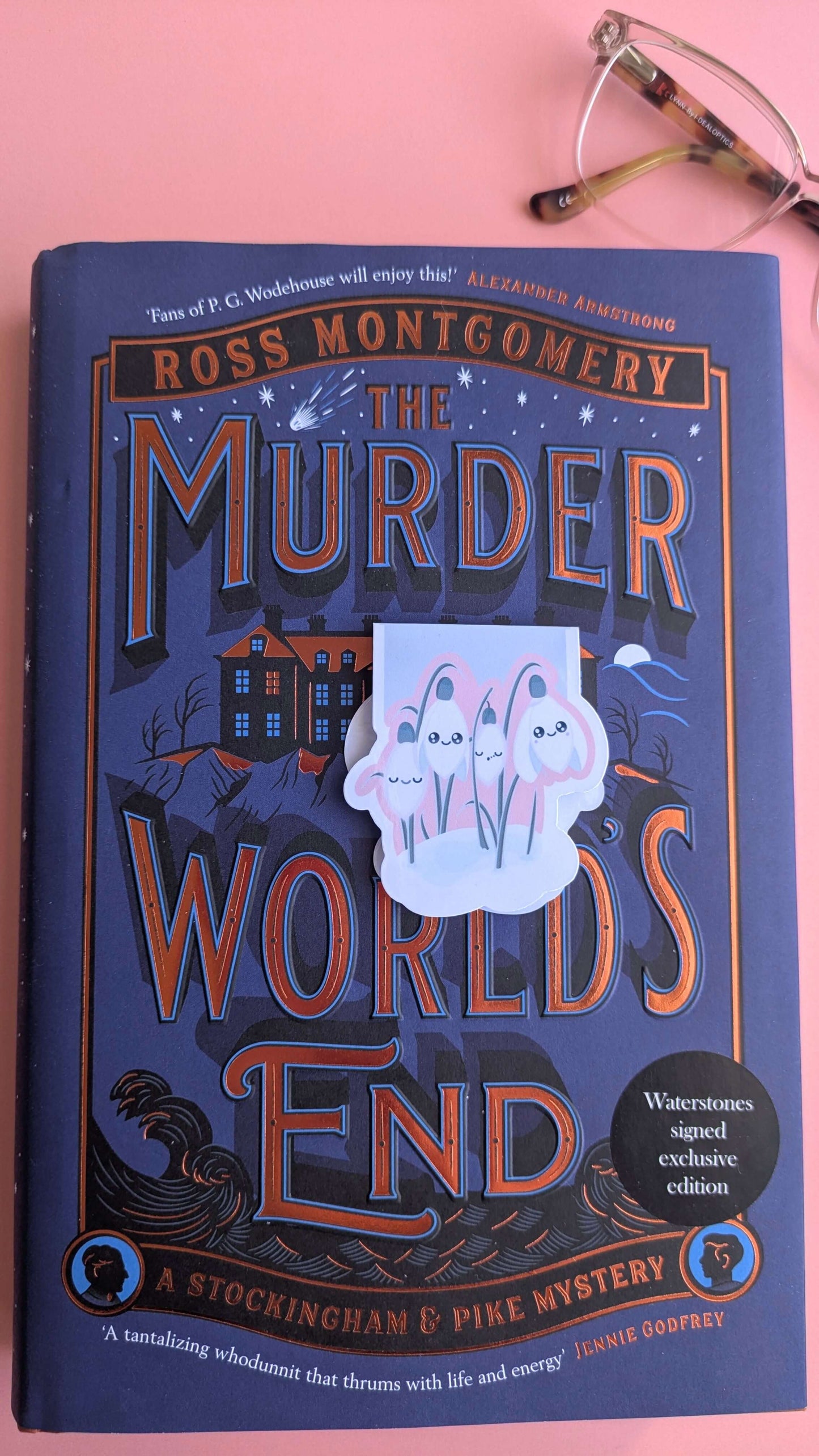 A Kawaii Sleepy Snowdrop 'Icy Venusian' pink magnetic bookmark placed centrally on 'The Murder at World’s End'. This minimalist composition removes the porcelain cup, focusing on the bookmark and reading glasses to showcase a clean, romanticized book-safe decorating style.