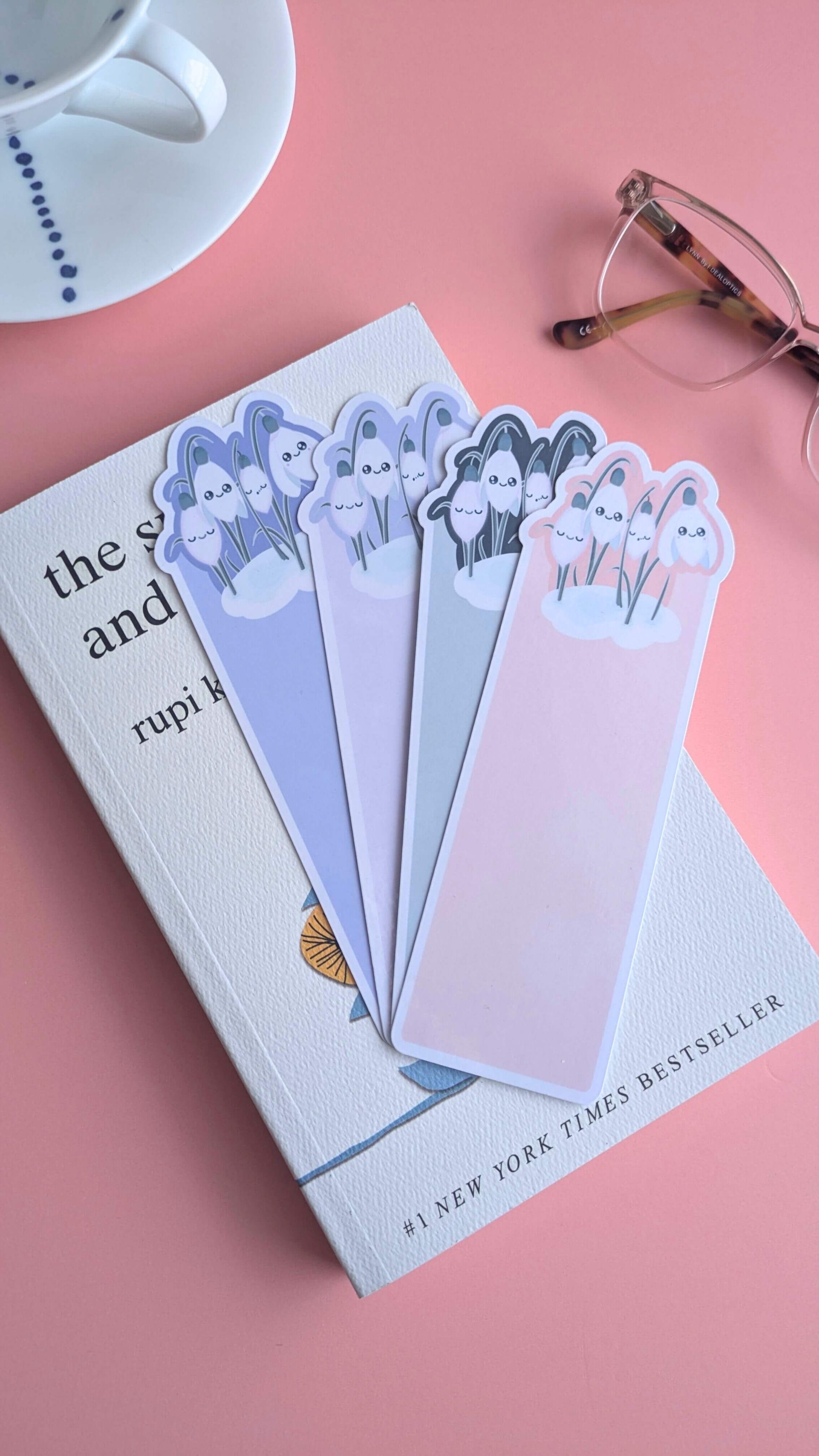 A fanned and slightly overlapping arrangement of the four Kawaii Sleepy Snowdrop bookmarks—Icy Venusian Pink, Dawn Lavender Blue, Midnight Mist, and Winter’s Edge Teal—on top of a closed copy of Rupi Kaur’s 'The Sun and Her Flowers'. The die-cut snowdrop clusters are centered in the frame, styled with a white porcelain saucer and reading glasses on a rose-pink background under soft morning window light to showcase the full "Obsessed with Color" collection.