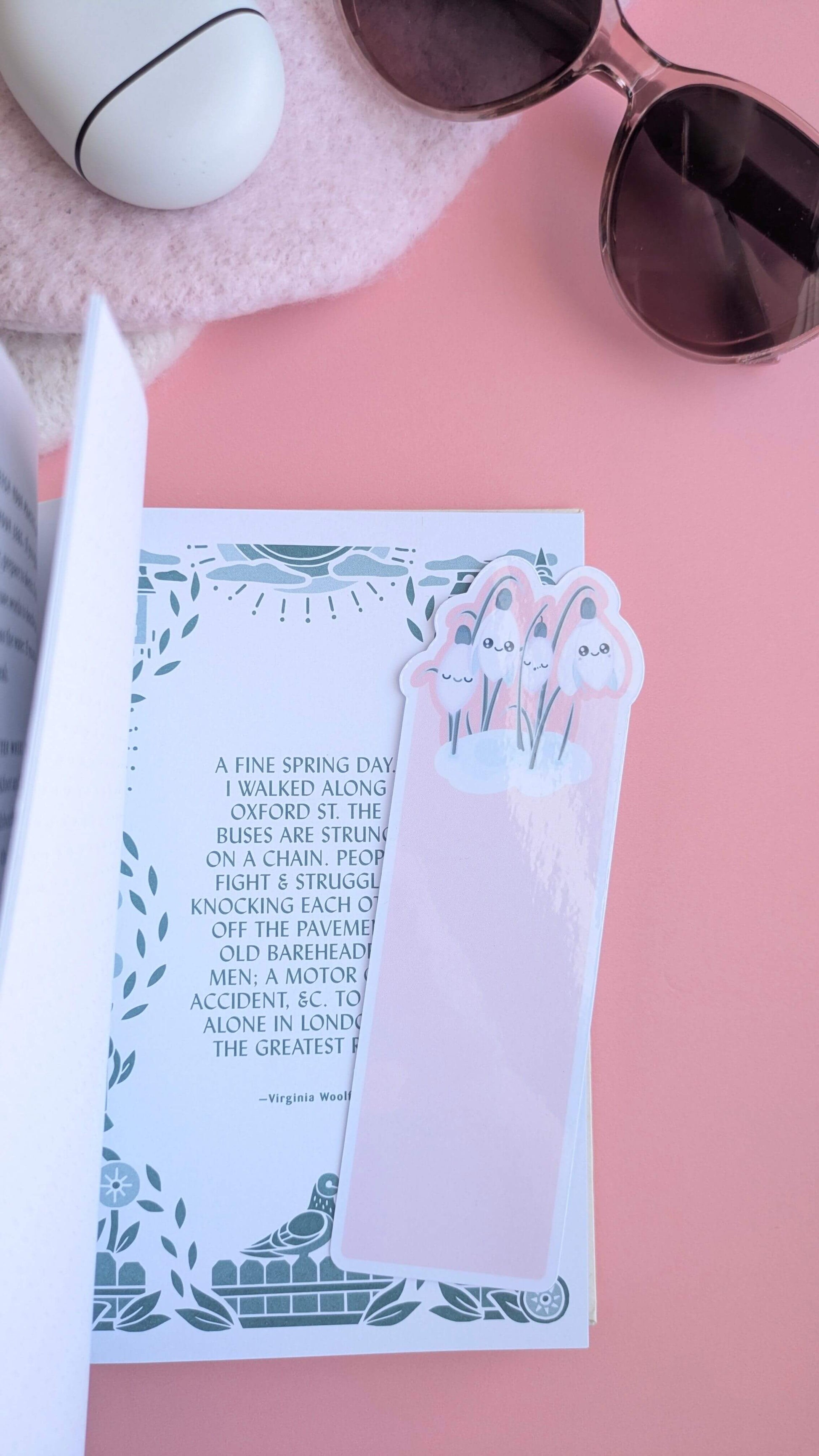 An Ateu Studio Kawaii Sleepy Snowdrop 'Icy Venusian Pink' die-cut bookmark placed on an open page of 'Afoot and Lighthearted: A Journal for Mindful Walking' which show a passage by Virginia Woolf. The silver-sage botanical illustrations and font color in the book perfectly mirror the bookmark’s sage-silver leaves. Styled with Fossil-style translucent sunglasses and a soft pink hat, this aerial view captures a mindful morning of reading and intentionality on a rose-pink background.