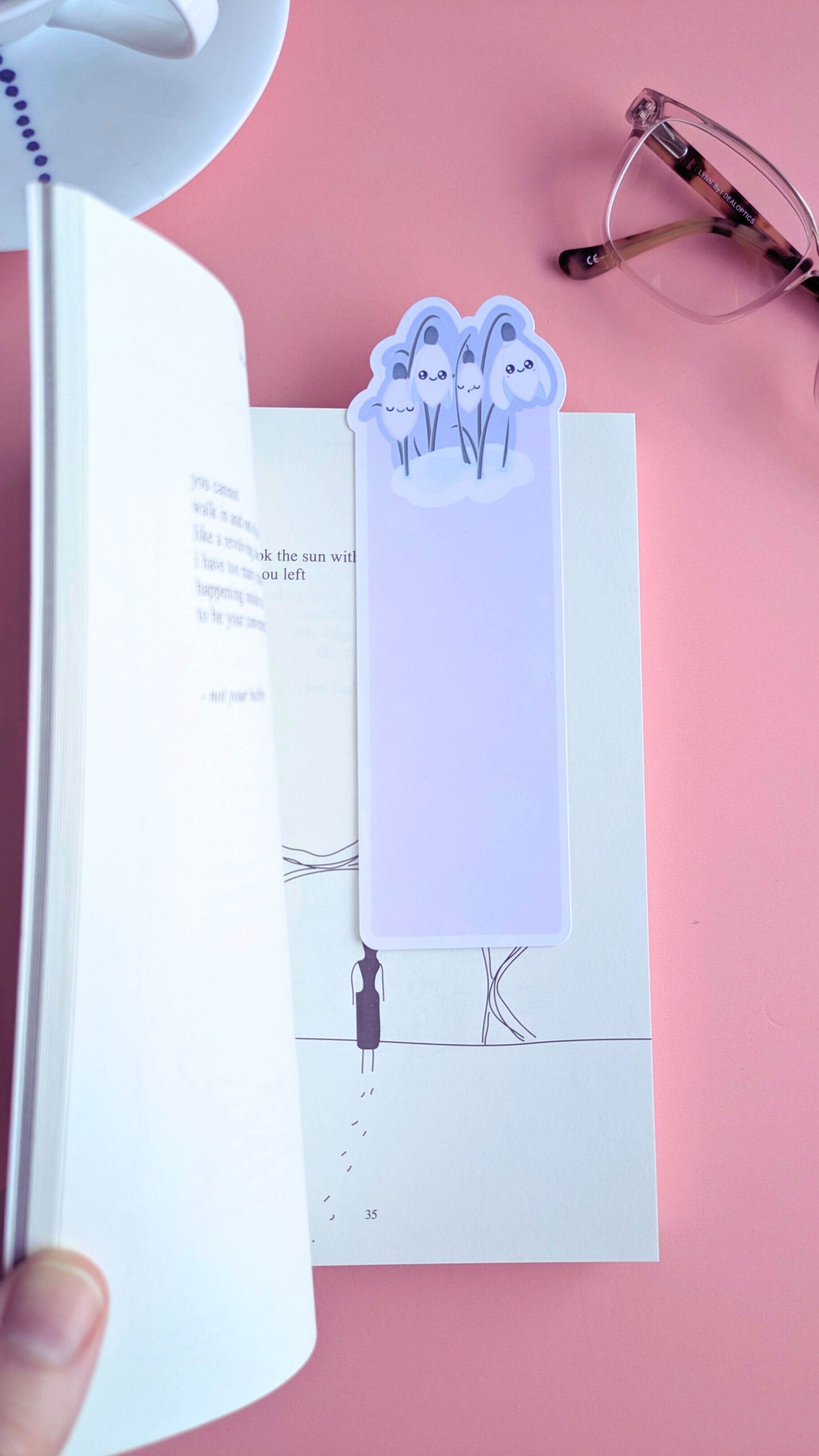 An Ateu Studio Kawaii Sleepy Snowdrop 'Dawn' lavender blue bookmark placed inside an open copy of 'The Sun and Her Flowers' by Rupi Kaur. The die-cut snowdrop silhouette and Snowdrop White characters contrast against the book pages, styled in an aerial view with a white porcelain cup and saucer and reading glasses. Soft morning window light on a rose-pink background highlights this mindful reading ritual and aesthetic book annotating setup.