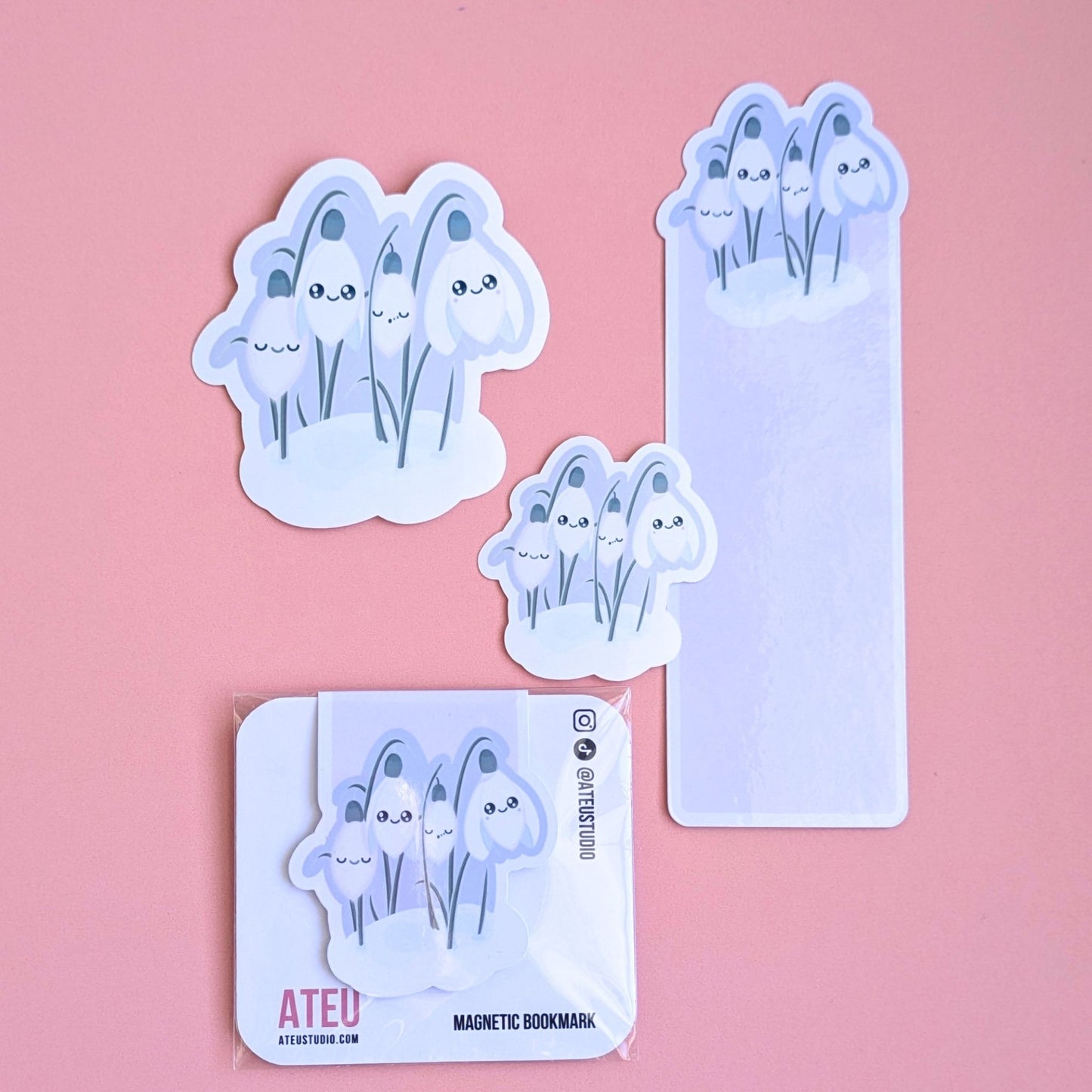 Ateu Studio Sleepy Snowdrop 'Dawn' Kawaii stationery gift set featuring Snowdrop White characters with sage-silver leaves and a cozy soft-glow effect. This lavender-blue and rose-pink aerial view showcases the die-cut bookmark, packaged magnetic bookmark, and two gloss vinyl stickers. Centered and clean, the layout highlights the high-shine gloss finish for Kindle decorating, book annotating, and gifting to aesthetic stationery lovers.