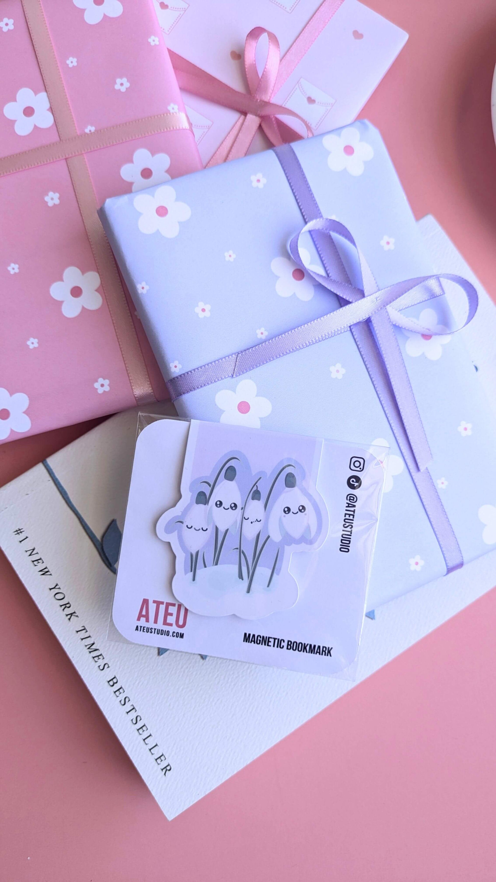 An Ateu Studio Kawaii Sleepy Snowdrop 'Dawn' Lavender Blue magnetic bookmark in retail packaging, styled as the centerpiece of a gift-giving flat lay. The composition includes stacked Ateu Studio daisy and envelope-patterned gifts, a copy of Rupi Kaur’s 'The Sun and Her Flowers' with the '#1 New York Times Best Seller' text visible, and a glimpse of a porcelain saucer. Soft natural light creates a premium gloss sheen on the bookmark packaging, perfect for aesthetic gift-giving inspiration.