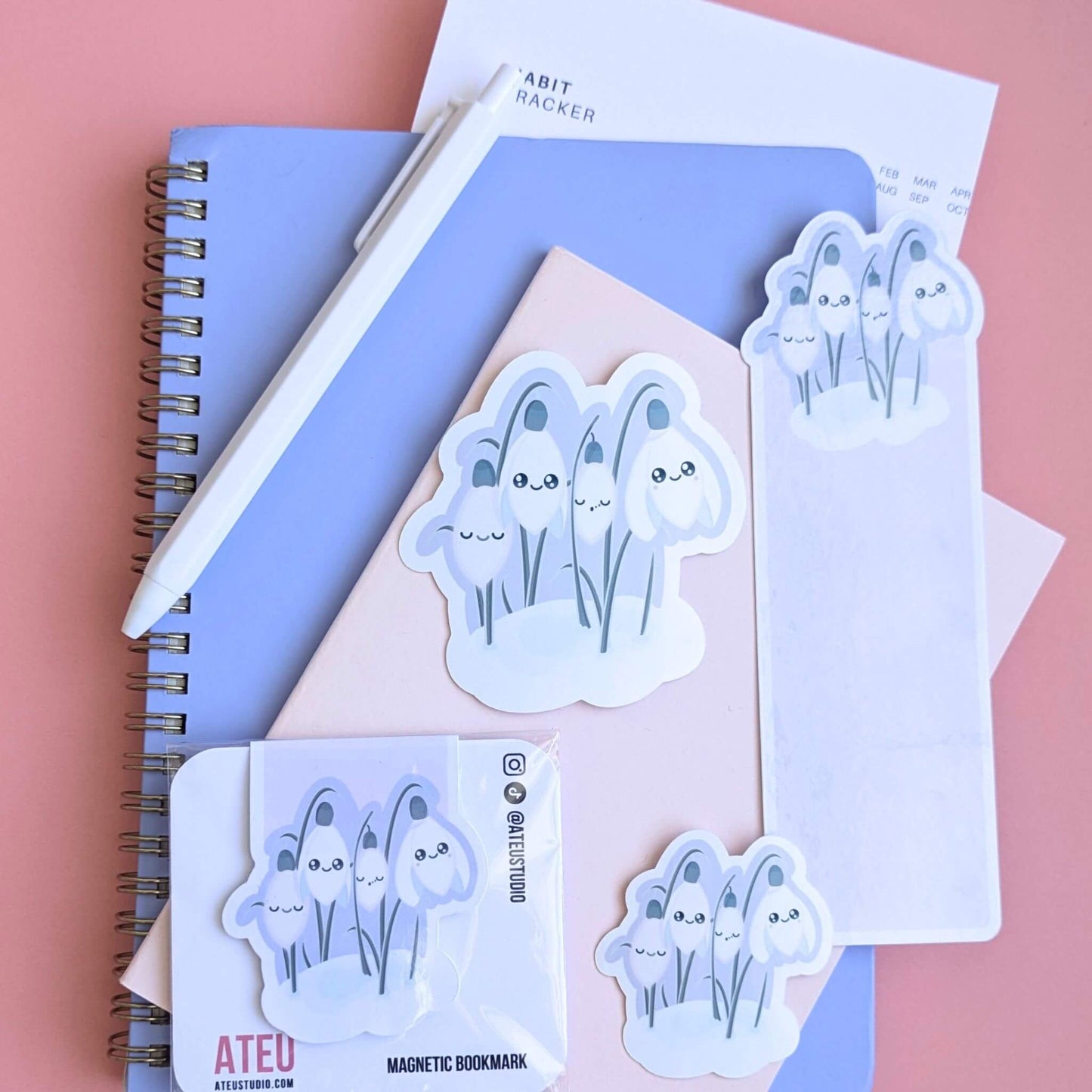 Ateu Studio Sleepy Snowdrop 'Dawn' stationery collection in a lavender-blue sky palette; Snowdrop White characters with sage-silver leaves and a cozy soft-glow effect. This aerial flat lay on a rose-pink background includes gloss vinyl stickers, a die-cut bookmark, and a packaged magnetic bookmark styled with a Taja spiral notebook, pink pocket journal, and habit tracker. This organized desk scene captures a mindful morning routine, intentional goal setting, book annotating, and aesthetic planning.
