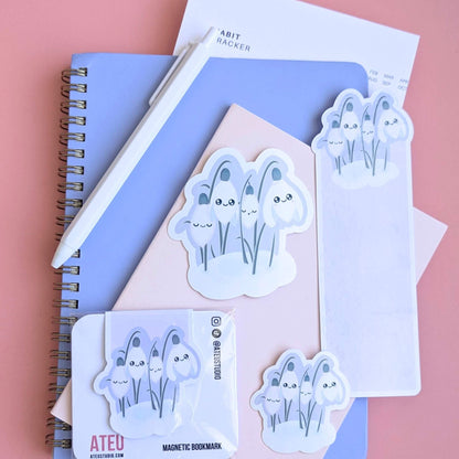 Ateu Studio Sleepy Snowdrop 'Dawn' stationery collection in a lavender-blue sky palette; Snowdrop White characters with sage-silver leaves and a cozy soft-glow effect. This aerial flat lay on a rose-pink background includes gloss vinyl stickers, a die-cut bookmark, and a packaged magnetic bookmark styled with a Taja spiral notebook, pink pocket journal, and habit tracker. This organized desk scene captures a mindful morning routine, intentional goal setting, book annotating, and aesthetic planning.