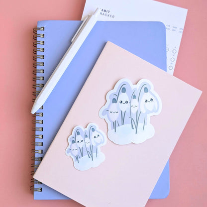 Ateu Studio Sleepy Snowdrop 'Dawn' 2-inch and 3-inch gloss vinyl stickers layered on a pink pocket notepad for size comparison. This lavender-blue and Snowdrop White character set is styled with a Taja spiral notebook, a habit tracker, and an off-white pen on a rose-pink background. The aerial flat lay highlights the soft-glow effect and sage-silver leaves, perfect for intentional goal setting and planner decorating.