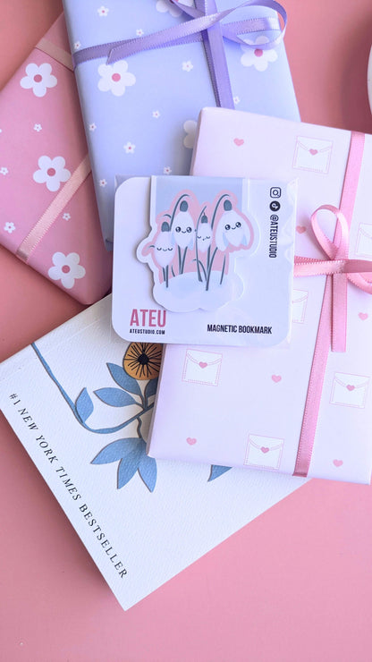 An Ateu Studio Kawaii Sleepy Snowdrop 'Icy Venusian' Pink magnetic bookmark in retail packaging, styled as the centerpiece of a gift-giving flat lay. The composition includes stacked Ateu Studio daisy and envelope-patterned gifts, a copy of Rupi Kaur’s 'The Sun and Her Flowers' with the '#1 New York Times Best Seller' text visible, and a glimpse of a porcelain saucer. Soft natural light creates a premium gloss sheen on the bookmark packaging, perfect for aesthetic gift-giving inspiration.