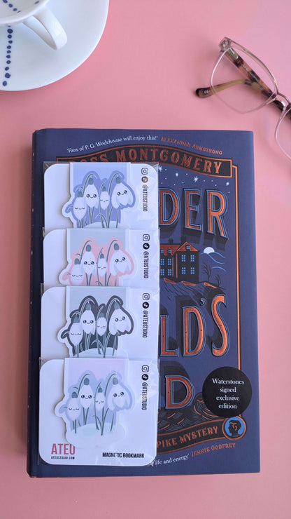 A vertical, zoomed-out aerial view of the full Ateu Studio Kawaii Sleepy Snowdrop magnetic bookmark collection. Four packaged variants are arranged vertically on the left of 'The Murder at World’s End' signed edition. The shot reveals reading glasses and the hand-painted blue dot detail inside the porcelain cup, creating a spacious, high-end "visual life-building" scene on a pink background.
