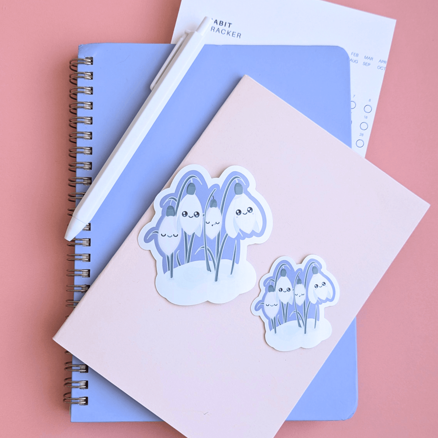 Ateu Studio Sleepy Snowdrop 'Midnight Mist' Lavender Blue stationery collection in a Ethereal Lavender sky palette. This aerial flat lay on a rose-pink background includes a gloss vinyl sticker, die-cut bookmark, and packaged magnetic bookmark styled with a Taja spiral notebook and habit tracker for a mindful morning routine and intentional goal setting.