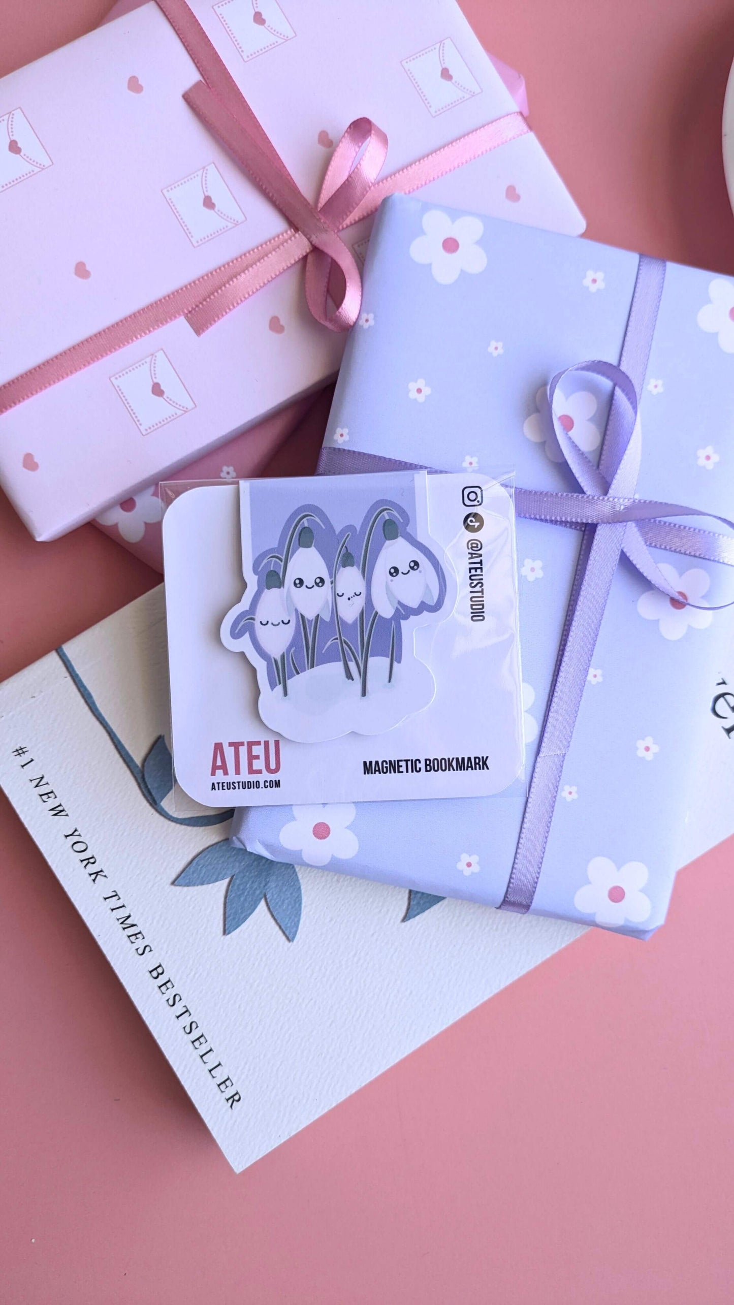 Aerial flat lay featuring the Ateu Studio Kawaii Sleepy Snowdrop 'Midnight Mist' magnetic bookmark in its retail packaging. Styled with three small, beautifully wrapped gifts: two stacked pink daisy-patterned packages tied with thin satin bows, and a pale pink envelope design with a heart seal. Rupi Kaur's 'The Sun and Her Flowers' sits as the central literary anchor. A soft light creates a premium sheen on the bookmark packaging, highlighting the high-quality presentation for an aesthetic gift giving setup