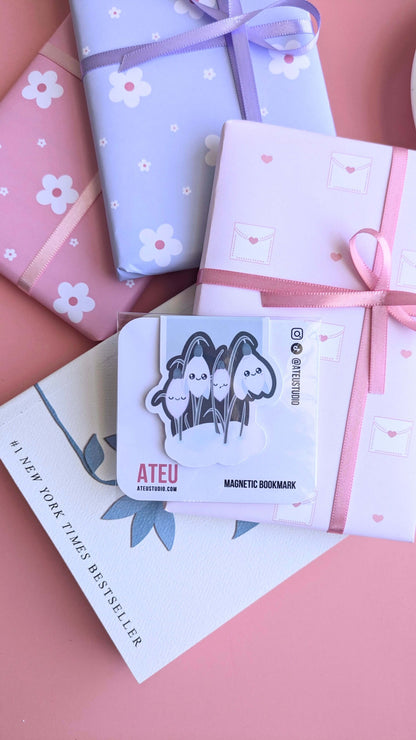 An Ateu Studio Kawaii Sleepy Snowdrop 'Winter’s Edge' Teal magnetic bookmark in retail packaging, styled as the centerpiece of a gift-giving flat lay. The composition includes stacked Ateu Studio daisy and envelope-patterned gifts, a copy of Rupi Kaur’s 'The Sun and Her Flowers' with the '#1 New York Times Best Seller' text visible, and a glimpse of a porcelain saucer. Soft natural light creates a premium gloss sheen on the bookmark packaging, perfect for aesthetic gift-giving inspiration.