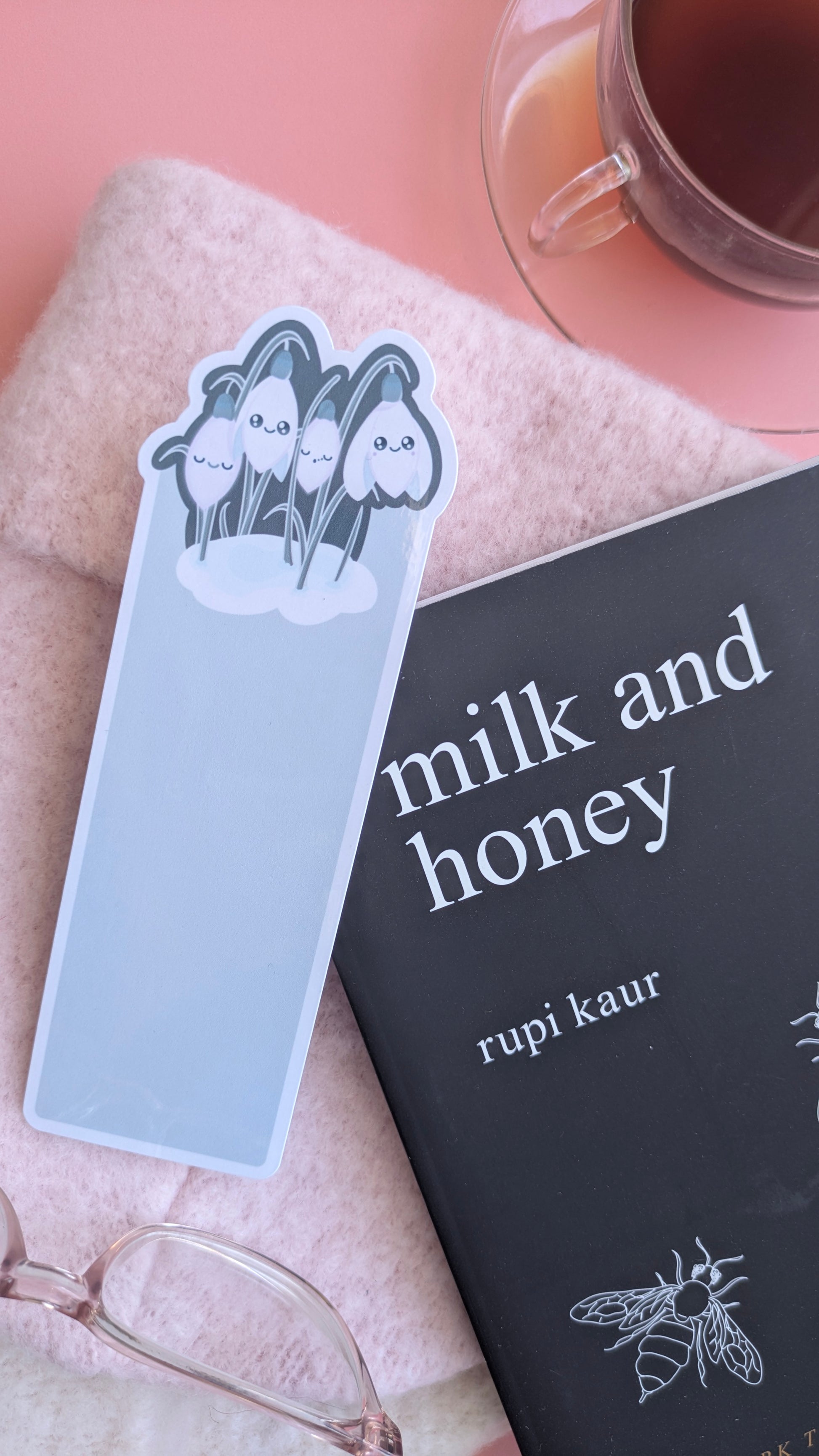 An Ateu Studio Kawaii Sleepy Snowdrop 'Winter’s Edge' teal bookmark angled across the lower quarter of a closed copy of Rupi Kaur’s 'Milk and Honey'. The black book cover with white bee illustration complements the darkest version of the teal collection. Styled on a soft, elegant pink wool hat with a glass French champagne-region cup of Earl Grey tea with honey and reading glasses, capturing a cozy winter reading ritual.