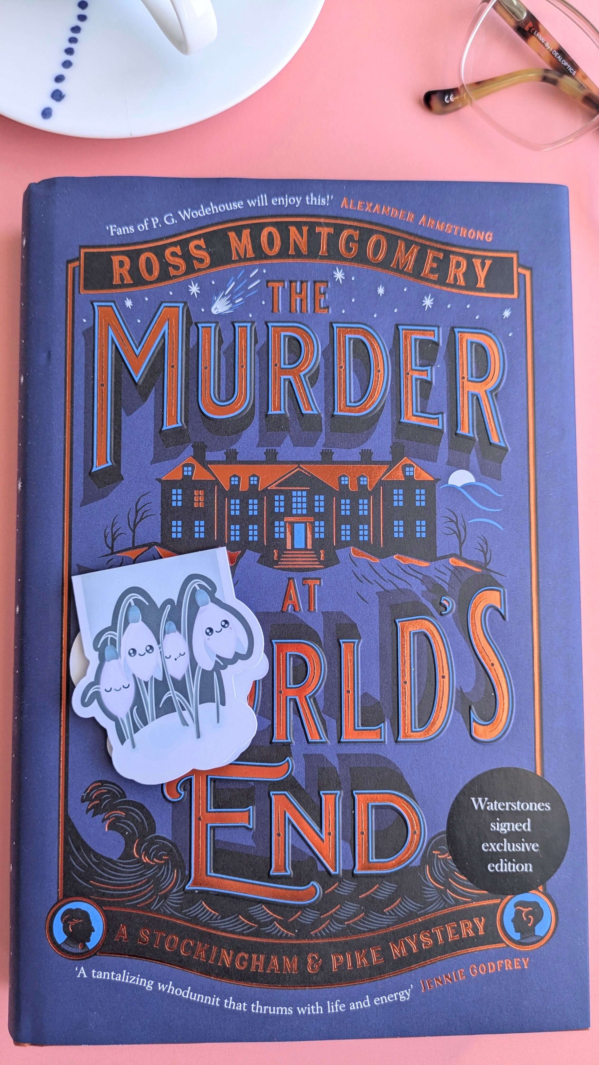 An unpackaged Kawaii Sleepy Snowdrop 'Winter’s Edge' teal magnetic bookmark resting on the lower left of a closed mystery novel. The unique die-cut shape is visible against the sprayed edges of the book, styled with a glass-rimmed porcelain saucer and reading glasses in a cozy winter reading scene.