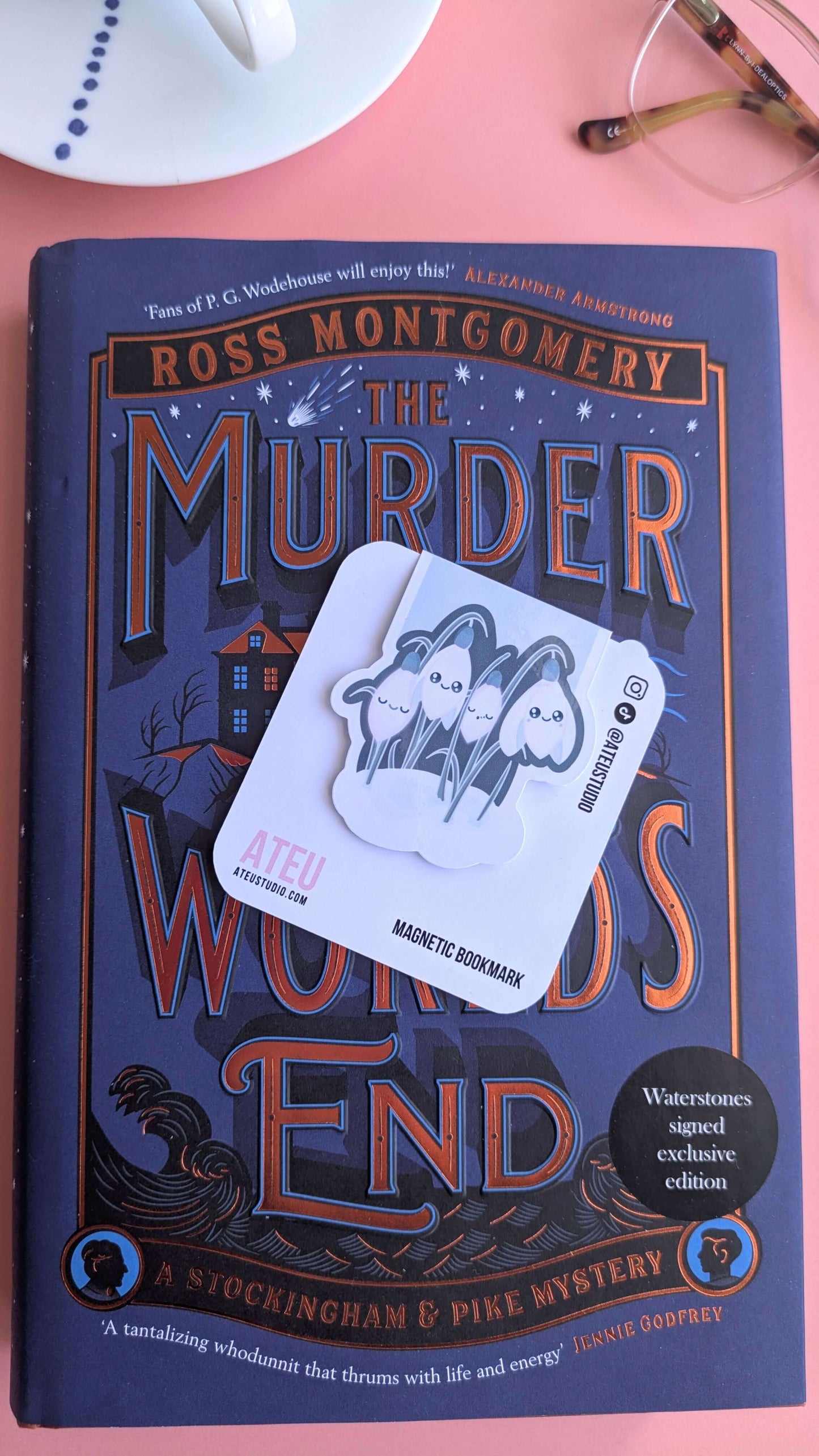 The Kawaii Sleepy Snowdrop 'Winter’s Edge' teal magnetic bookmark on its branded card, offset to the right to draw out the deep blue and bronze tones of the Ross Montgomery book cover. Styled with a French porcelain saucer and reading glasses, highlighting the darker teal palette against the celestial book design.