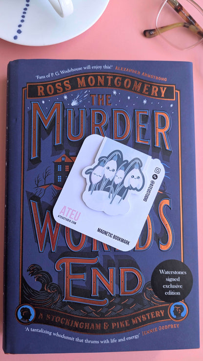 The Kawaii Sleepy Snowdrop 'Winter’s Edge' teal magnetic bookmark on its branded card, offset to the right to draw out the deep blue and bronze tones of the Ross Montgomery book cover. Styled with a French porcelain saucer and reading glasses, highlighting the darker teal palette against the celestial book design.