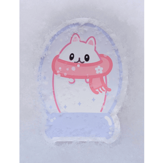 Whimsical lifestyle photo of the Lil Beta winter snowglobe sticker placed in real snow during Winter Storm Fern. Real snowflakes rest on the gloss vinyl surface, highlighting the lavender and pink colors of the egg-shaped cat design.