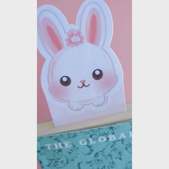 A close-up video of the Marshmallow Blush Bunny Pink bookmark tucked into a closed book. The camera tilts to catch the natural window light, revealing a premium high-gloss sheen on the white and pink character design. This Ateu Studio accessory elevates any reading aesthetic through vibrant color and quality finish.