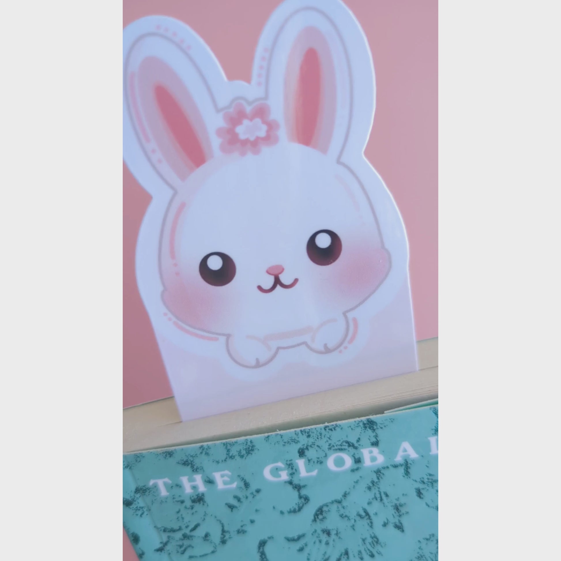 A close-up video of the Marshmallow Blush Bunny Pink bookmark tucked into a closed book. The camera tilts to catch the natural window light, revealing a premium high-gloss sheen on the white and pink character design. This Ateu Studio accessory elevates any reading aesthetic through vibrant color and quality finish.