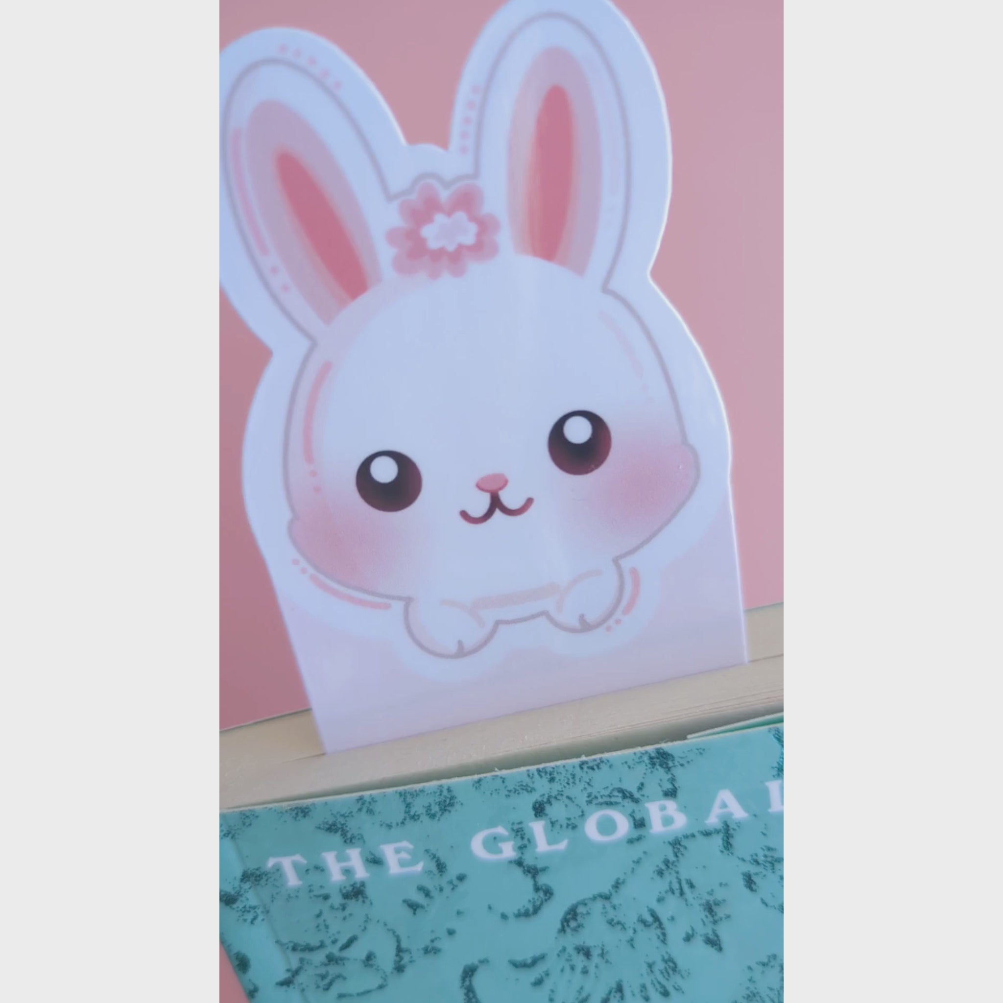 A close-up video of the Marshmallow Blush Bunny Pink bookmark tucked into a closed book. The camera tilts to catch the natural window light, revealing a premium high-gloss sheen on the white and pink character design. This Ateu Studio accessory elevates any reading aesthetic through vibrant color and quality finish.