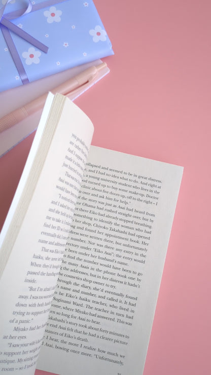 A point-of-view video showing a hand placing the Daffodil Hug Szn 'Sunny Shine' yellow magnetic bookmark onto a book page and gently 'booping' it closed with an index finger. The video highlights the satisfying magnetic snap and secure grip of the gloss-finish bookmark before the book is closed. The coordinated daisy gift wrap and pink stationery in the background reinforce the Ateu Studio aesthetic for mindful reading rituals.