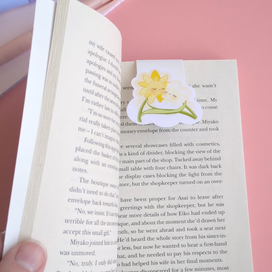 A short, point of view video demonstrating the Daffodil Hug Szn 'Daffodil White' magnetic bookmark. A thumb holds the book page open, revealing the bookmark in place with the magnets separated. An index finger gently 'boops' the character, snapping the strong magnetic clasp closed on the page. The coordinated daisy gift wrap and pink notepad in the background reinforce the curated reading sanctuary.
