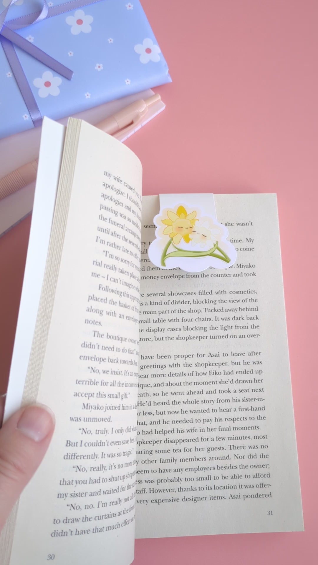 A short, point of view video demonstrating the Daffodil Hug Szn 'Daffodil White' magnetic bookmark. A thumb holds the book page open, revealing the bookmark in place with the magnets separated. An index finger gently 'boops' the character, snapping the strong magnetic clasp closed on the page. The coordinated daisy gift wrap and pink notepad in the background reinforce the curated reading sanctuary.