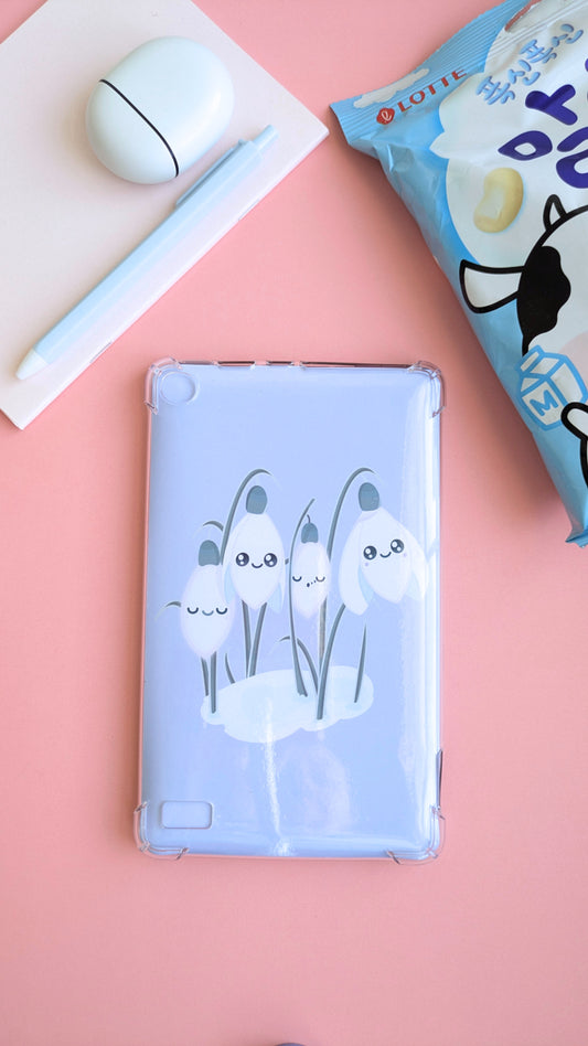A full aerial desk-scape featuring the 'Sleepy Snowdrops Dawn Lavender Blue Kindle Paperwhite Insert' inside a clear case. The composition includes a pink notepad with Pixel Buds, a white pen, a fine porcelain saucer, and a packet of Lotte Malang Cow Chewy Candy for a fresh winter aesthetic.