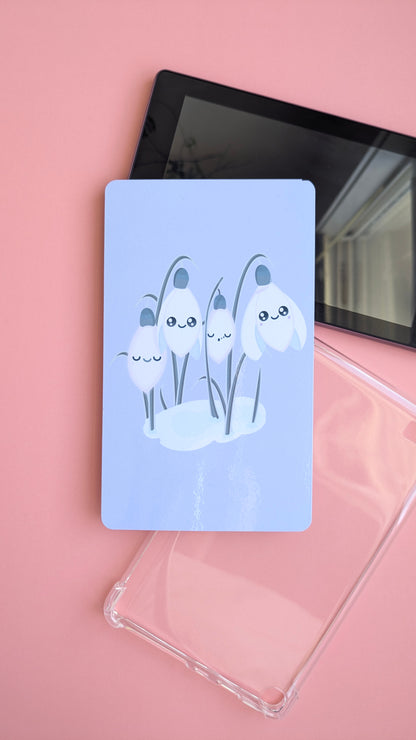 A deconstructed view of the 'Sleepy Snowdrops Dawn Lavender Blue Kindle Paperwhite Insert' propped against a clear case. This shot highlights the heavy gloss sheen and a natural window reflection with jasmine plants, showing the adhesive-free setup for 6th, 10th, 11th and 12th Gen models.