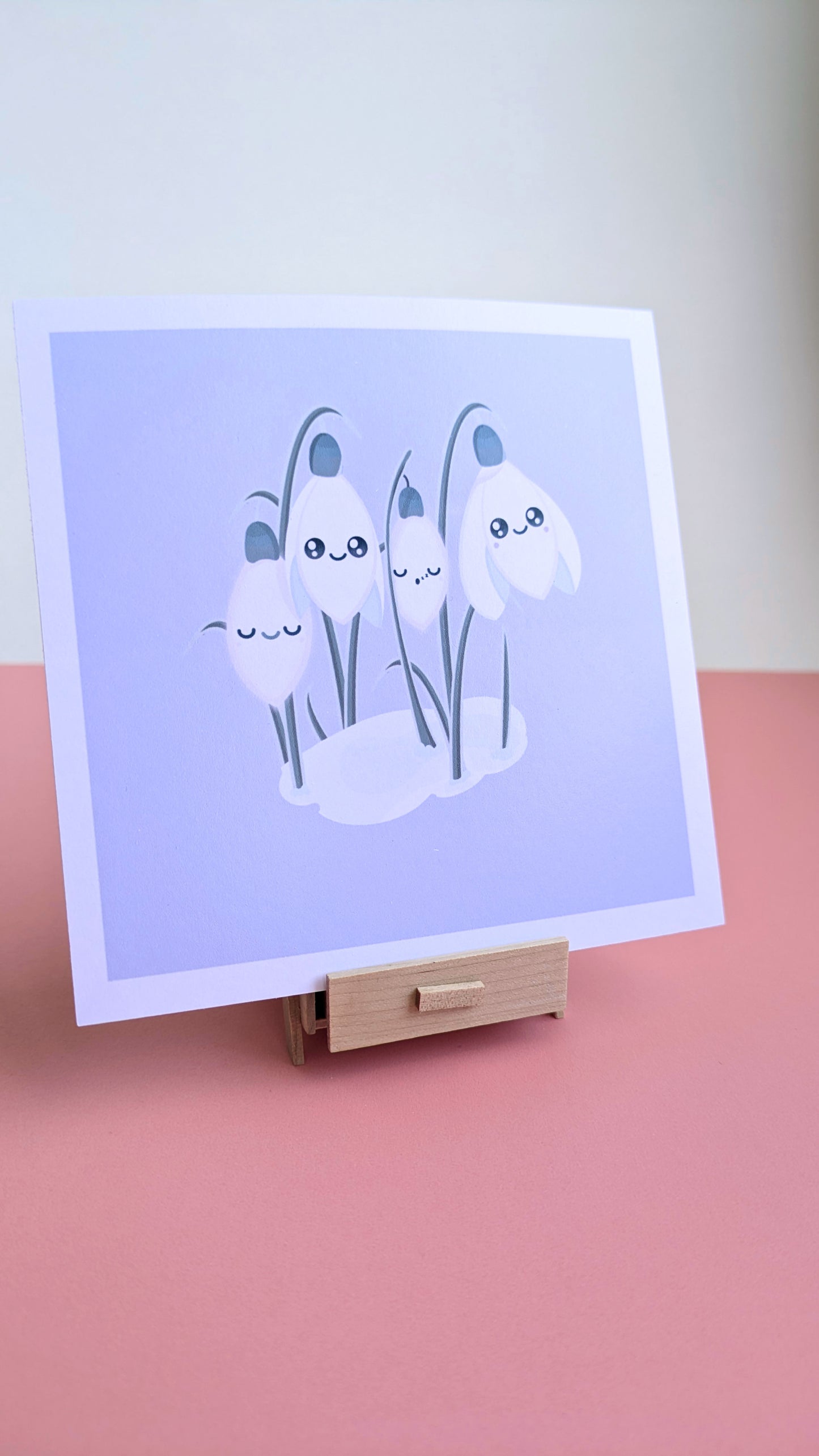A 5x5 'Sleepy Snowdrops Dawn Lavender Blue Mini Art Print' on a pink desk with a white wall behind it. The original kawaii artwork is positioned on a miniature wooden stand, showing the smooth, low glare finish and clean white border in natural light for a fresh winter desk reset.