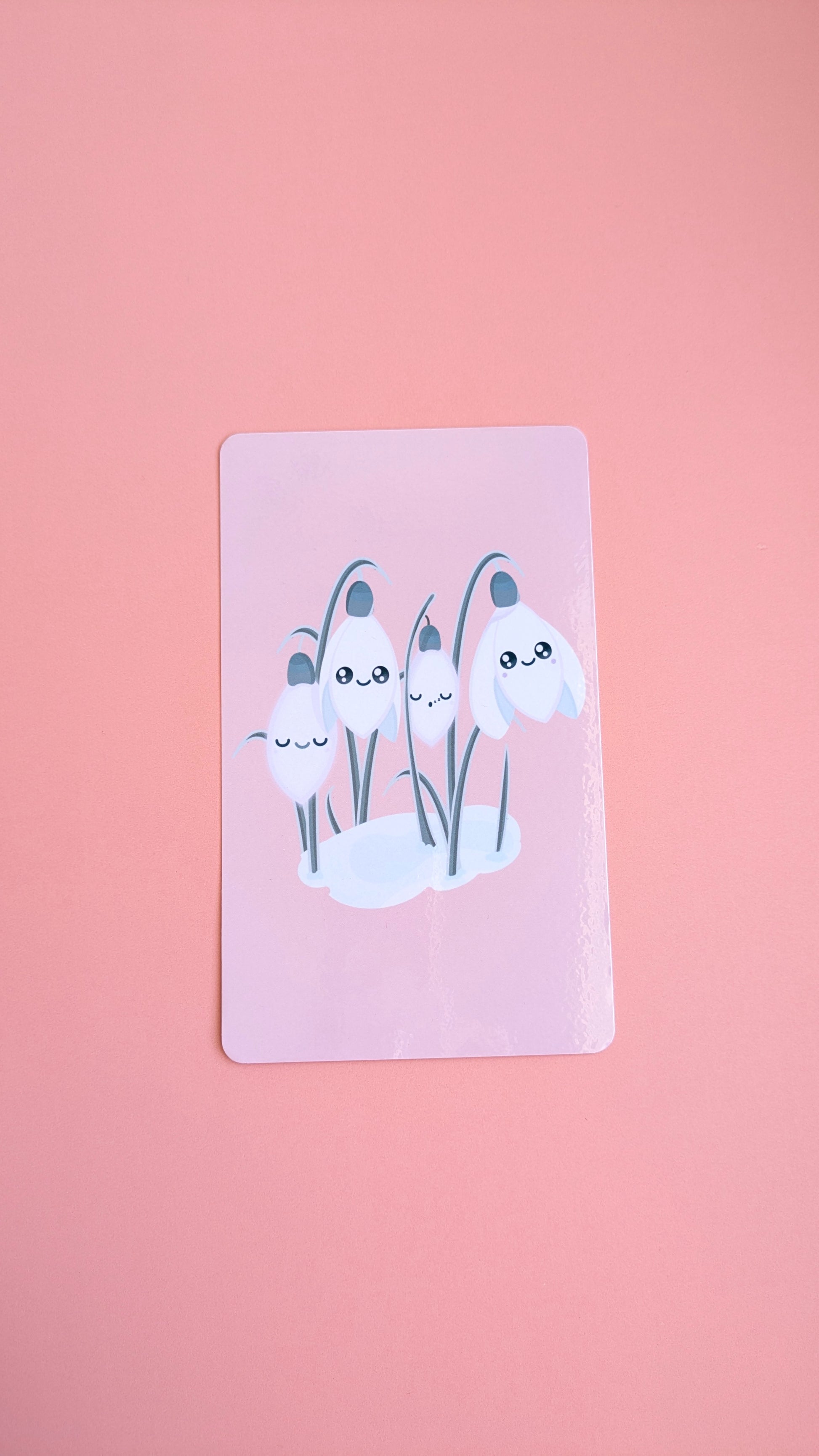 An aerial, naturally lit view of the 'Sleepy Snowdrops Icy Venusian Pink Kindle Paperwhite Insert' on a pink background. The high-res photo media features the original kawaii characters against an icy blush celestial sky with a high-gloss finish.