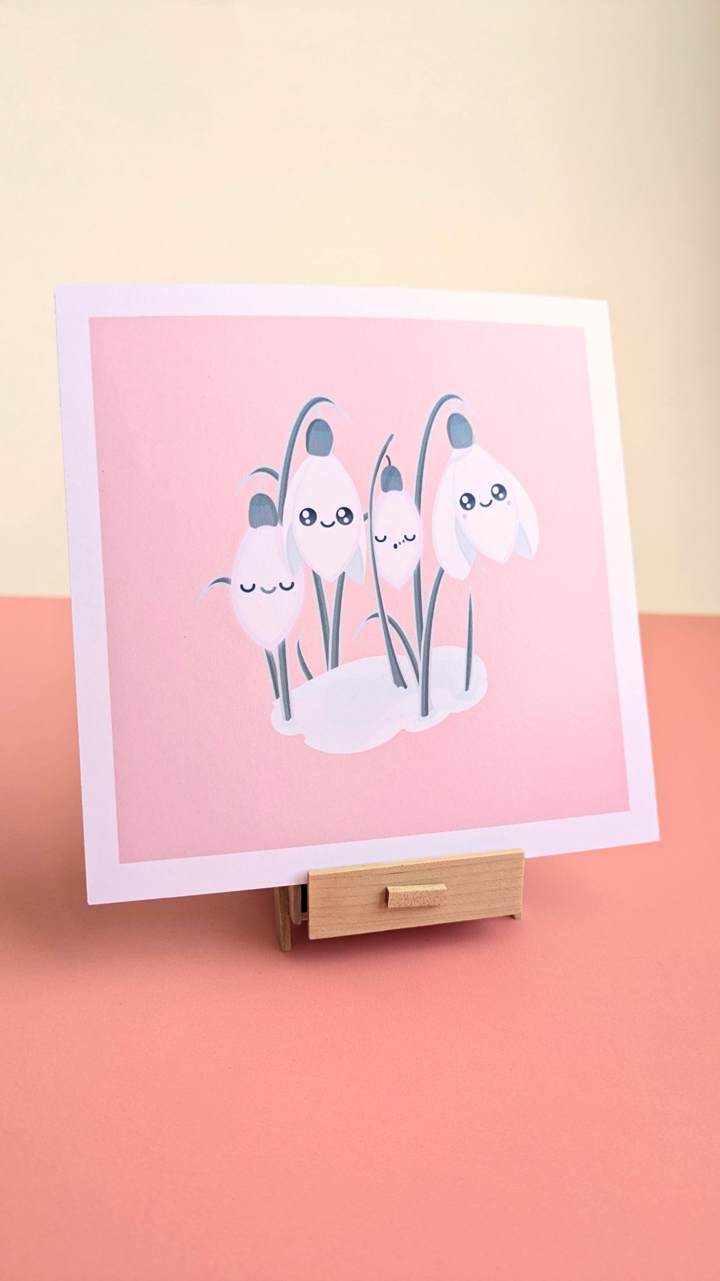 A 5x5 'Sleepy Snowdrops Icy Venusian Pink Mini Art Print' on a pink desk with a white wall behind it. The hand-drawn celestial artwork, featuring original kawaii characters against an icy blush sky, rests on a subtle miniature wooden stand. Natural light highlights the smooth, low-glare finish and clean white border, creating soft shadows for a curated winter desk aesthetic.