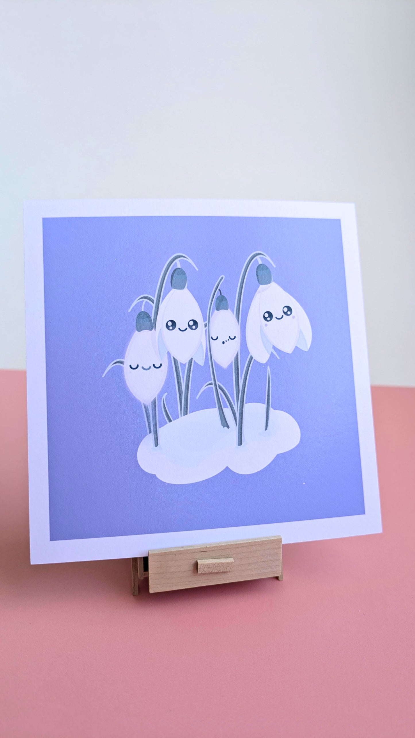 A centered 5x5 'Sleepy Snowdrops Midnight Mist Lavender Blue Mini Art Print' on a pink desk with a white wall behind it. The artwork is resting on a miniature wooden stand, highlighting the lavender-blue sky and original kawaii characters in a reflection-free, natural light composition.