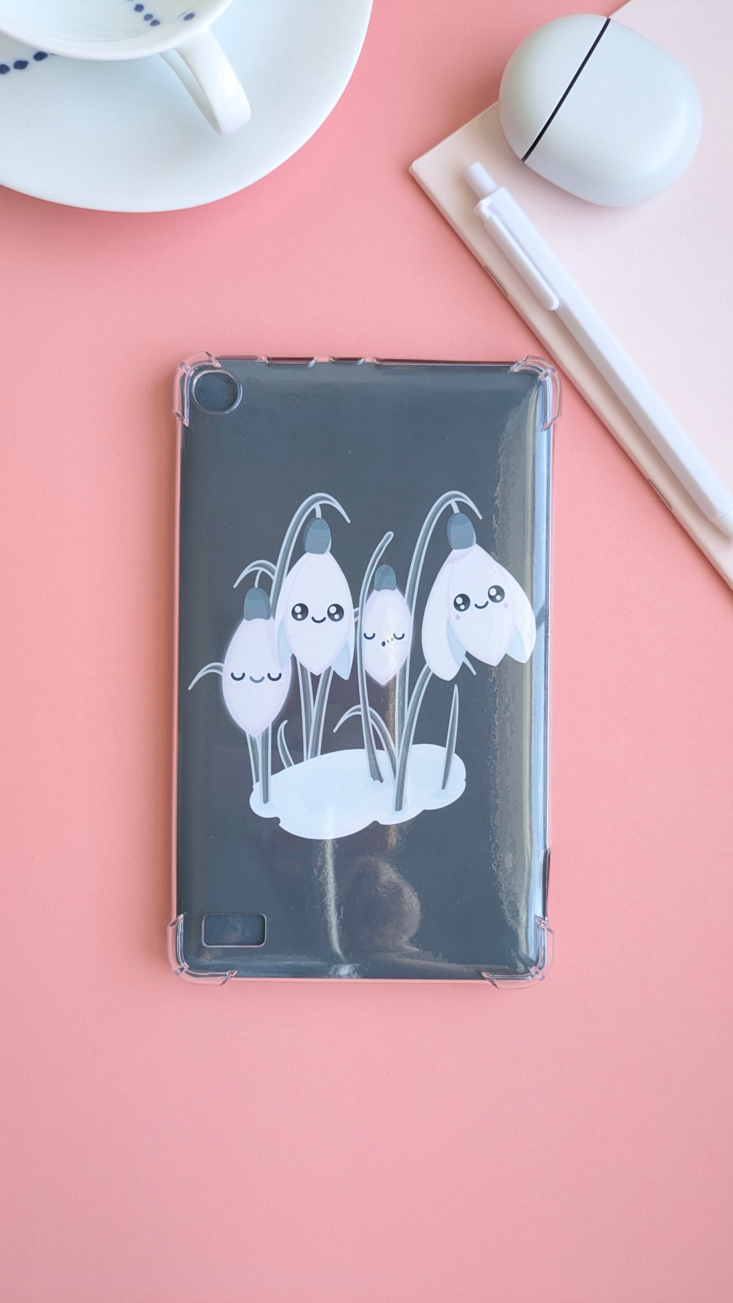 An aerial desk-scape featuring the 'Sleepy Snowdrops Winter’s Edge Teal Kindle Paperwhite Insert' inside a clear case. The composition includes a pink notepad, Pixel Buds, and a fine porcelain cup with hand-painted blue dots, highlighting a heavy gloss sheen and coordinated winter desk reset.