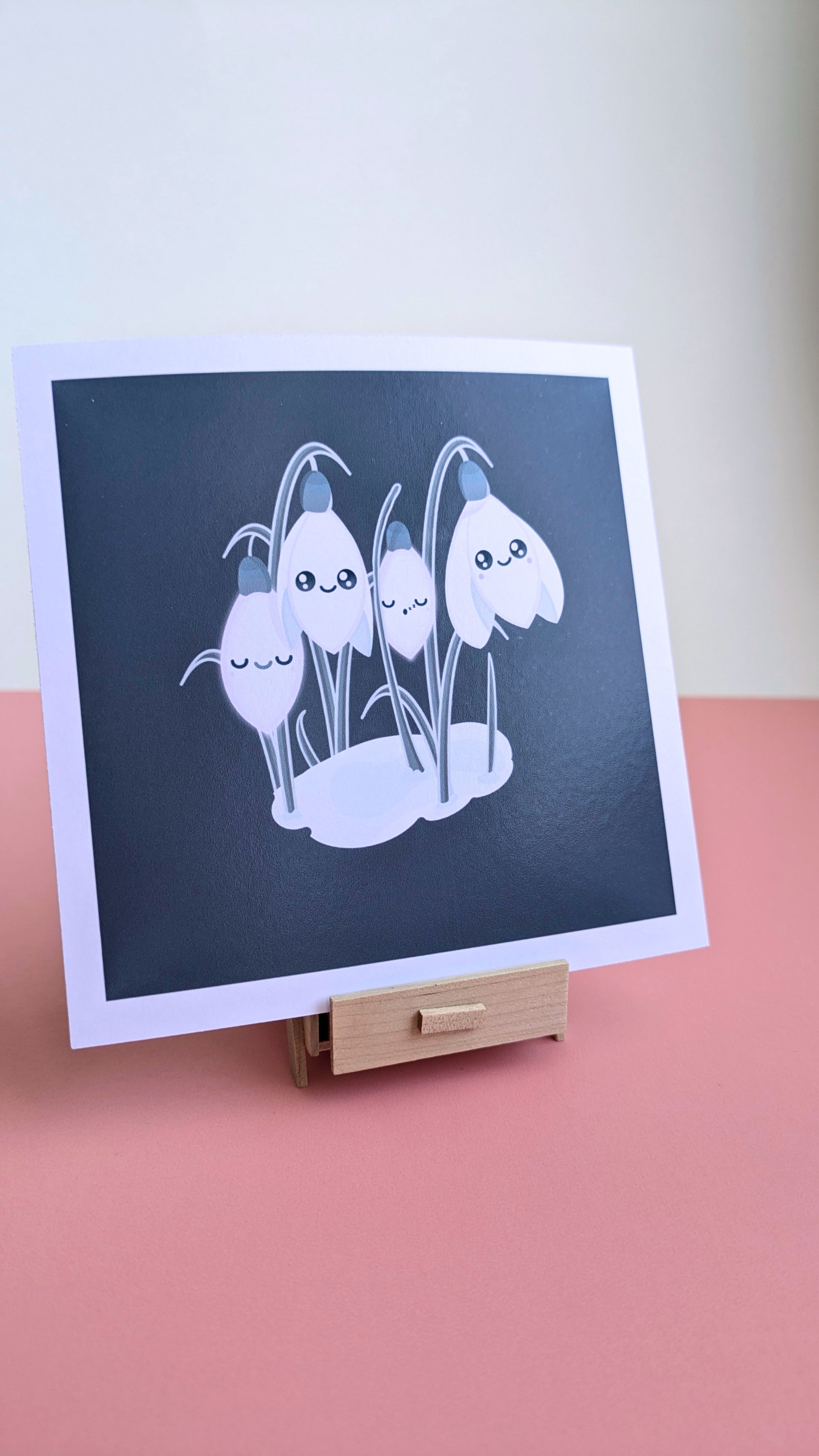 A 5x5 'Sleepy Snowdrops Winter’s Edge Teal Mini Art Print' on a pink desk with a white wall behind it. The deep teal winter sky and kawaii character design are featured on a miniature wooden stand, emphasizing the bold color contrast and smooth finish for a curated winter desk reset.